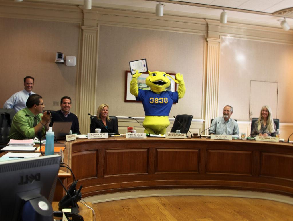 Sammy the mascot in the chambers of the Santa Cruz City council holding their hands overhead in celebration.